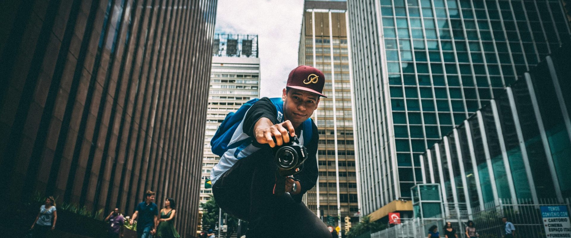 A young man with a camera kneels on a bustling city street, surrounded by skyscrapers.